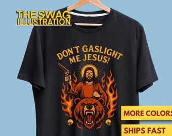 Könnte beinhalten: Schwarzes T-Shirt mit einer Grafik von Jesus mit einer Pistole, Flammen und einem Bären. Der Text lautet "DON'T GASLIGHT ME JESUS!". Das Logo The Swag Illustration befindet sich oben.