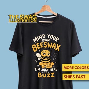 Puede incluir: Una camiseta negra de manga corta con un gráfico de una abeja gruñona con los brazos cruzados sentada sobre un panal. El texto dice "MIND YOUR OWN BEESWAX" e "I'M JUST HERE TO BUZZ" en letras amarillas y blancas, colgada en una percha de madera.