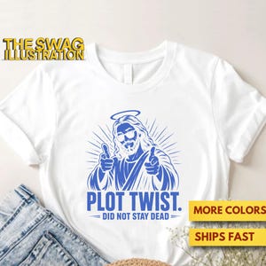 May include: A white short-sleeved t-shirt featuring a blue graphic of a figure with a halo and sunglasses pointing forward with both hands. Below the graphic, the text reads PLOT TWIST. DID NOT STAY DEAD. The shirt is displayed with blue denim.