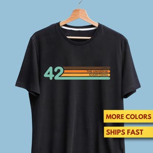 May include: Black t-shirt with the number "42" in teal and horizontal stripes in orange, brown, and teal. The text "LIFE THE UNIVERSE EVERYTHING" is below the stripes. The shirt is hanging on a wooden hanger.