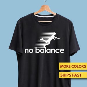 May include: Black t-shirt with the words "no balance" in white, and a white graphic of a running person. The shirt is displayed on a wooden hanger against a light blue backdrop.