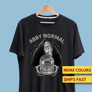 Puede incluir: Camiseta negra con una ilustración en escala de grises de un personaje sosteniendo un cerebro en una cúpula de cristal. El texto "ABBY NORMAL" está arqueado sobre el personaje. Debajo de la cúpula, el texto dice "OR USE THIS BRAIN AB-NORMAL".