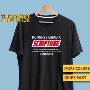 May include: Black t-shirt with the text "HUNGRY? GRAB A SCRIPTURE" in white and red. The shirt also features the Bible verse Matthew 5:6. The shirt is hanging on a wooden hanger.