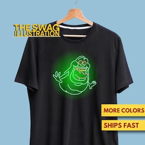 May include: Black t-shirt featuring a neon green cartoon character design. The design is a glowing outline of a smiling, blob-like figure. The text "THE SWAG ILLUSTRATION" is at the top. Additional text on the shirt says "MORE COLORS" and "SHIPS FAST".