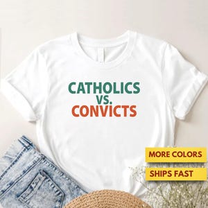 Puede incluir: Camiseta blanca con el texto "CATHOLICS vs. CONVICTS" en verde y naranja. La camiseta se combina con vaqueros azules y un sombrero de paja. El texto adicional en la imagen dice "MORE COLORS" y "SHIPS FAST".