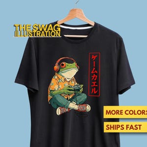 May include: Black t-shirt featuring a cartoon frog wearing headphones and playing a video game. The frog is dressed in a Hawaiian shirt and jeans. Japanese text is on the right side of the shirt. The Swag Illustration logo is at the top.