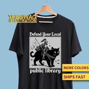 Op de afbeelding: Zwart T-shirt met een afbeelding van een ridder op een zwarte kat, met de tekst "Defend Your Local Public Library". De ridder draagt een vlag. Het shirt heeft een geel en wit logo met de tekst "THE SWAG ILLUSTRATION."