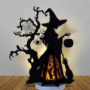 May include: Black silhouette of a witch holding a pumpkin, with a tree, spider web, and a cat inside a lantern. The Halloween decoration is illuminated from within, creating a spooky ambiance.