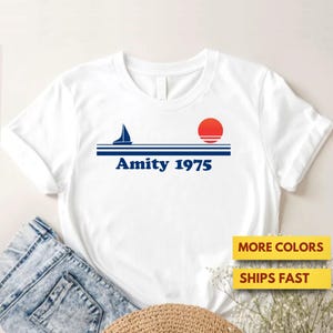 May include: White t-shirt featuring a nautical design with a sailboat, a red sun, and the text "Amity 1975" in blue. The shirt has a crew neck and short sleeves. The image also shows a pair of blue jeans and a straw hat.