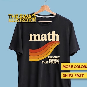 May include: Black t-shirt with the word "math" in cream-colored block letters and a retro orange and yellow striped design. The text "THE ONLY SUBJECT THAT COUNTS" is below the design. The shirt is hanging on a wooden hanger.