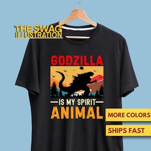 May include: Black t-shirt featuring a Godzilla graphic with the text "GODZILLA - IS MY SPIRIT - ANIMAL". The design includes a silhouette of Godzilla against a sunset backdrop. The shirt is displayed on a wooden hanger.