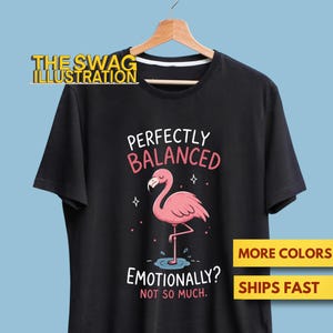 May include: A black short-sleeve t-shirt featuring a graphic of a pink flamingo standing on one leg in a small puddle. The text reads "PERFECTLY BALANCED" in white and pink, and "EMOTIONALLY? NOT SO MUCH." in white and red lettering.