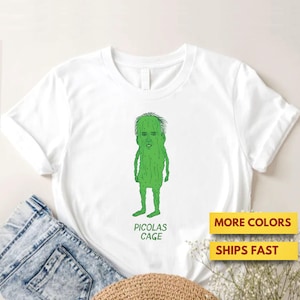 Puede incluir: Camiseta blanca con una ilustración de dibujos animados verde de un pepinillo con la cara de Nicolas Cage. El texto "PICOLAS CAGE" está debajo de la imagen. El texto adicional en la imagen dice "MORE COLORS" y "SHIPS FAST."