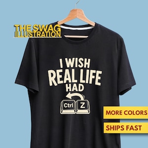 May include: A black short-sleeve t-shirt featuring the cream-coloured text I WISH REAL LIFE HAD followed by a graphic of two computer keyboard keys labelled Ctrl and Z with a curved arrow connecting them, all displayed on a wooden hanger.