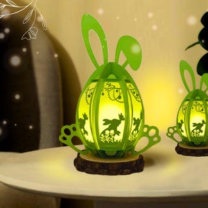 May include: Two green Easter egg-shaped lanterns with bunny ear accents. The lanterns have cut-out designs of bunnies and foliage, sitting on wooden bases. The lanterns are illuminated, creating a warm glow, and are placed on a white table.
