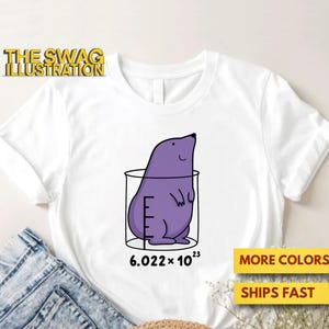 Op de afbeelding: Wit T-shirt met een paarse cartoon mol in een beker, met het getal 6,022 x 10^23. De tekst "THE SWAG ILLUSTRATION" staat linksboven. Extra tekst rechtsonder luidt "MORE COLORS" en "SHIPS FAST".