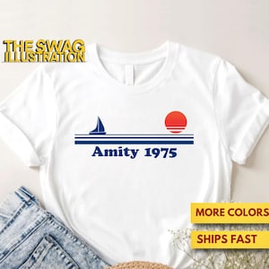 Könnte beinhalten: Weißes T-Shirt mit einem blauen Segelboot, einer roten Sonne und blauen Streifen. Der Text "Amity 1975" ist in Blau gedruckt.