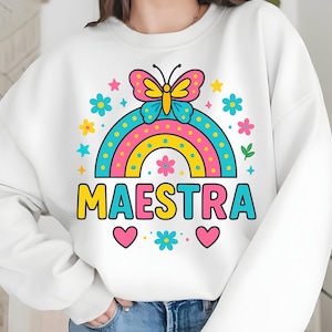 Puede incluir: Sudadera blanca con un gr&aacute;fico colorido. El dise&ntilde;o incluye un arco&iacute;ris, una mariposa, flores, estrellas y la palabra "MAESTRA" en una fuente divertida. El tema general es alegre y educativo.