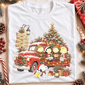 May include: A white t-shirt featuring a graphic of a red vintage truck loaded with a Christmas tree and gifts. Four cartoon characters in winter attire sit in the truck bed. A signpost reads Cocoa, Sleigh Rides, Christmas Trees, North Pole.