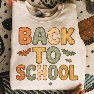 Könnte beinhalten: Beiges Sweatshirt mit dem Schriftzug "BACK TO SCHOOL" in Patchwork-Buchstaben. Die Buchstaben sind orange, grün und gelb mit Fledermaus- und Sternmotiven. Das Sweatshirt ist gefaltet.