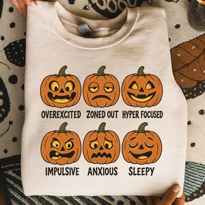 Op de afbeelding: Een beige sweatshirt met zes oranje cartoonpompoenen, elk met een andere gezichtsuitdrukking. De tekst onder elke pompoen luidt: overstuur, afwezig, hypergefocust, impulsief, angstig en slaperig.