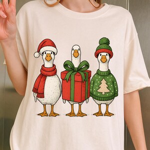 Könnte beinhalten: Cremefarbenes T-Shirt mit drei Cartoon-Gänsen in festlicher Kleidung. Eine Gans trägt einen Weihnachtsmannhut und einen roten Schal, eine andere hält ein Geschenk, und die dritte hat einen grünen Pullover mit einem Weihnachtsbaum-Design. Die Gänse stehen nebeneinander.