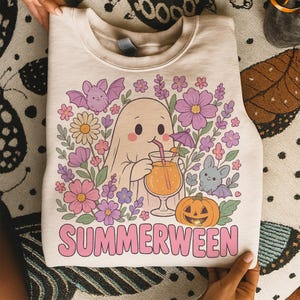May include: Cream-coloured sweatshirt with a cartoon ghost drinking a cocktail, surrounded by flowers, bats, and a jack-o'-lantern. The word "SUMMERWEEN" is printed in pink. A fun, seasonal design.
