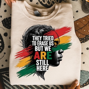 Op de afbeelding: Beige sweatshirt met een zwart-wit portret en rode, gele en groene penseelstreken. De tekst luidt: "THEY TRIED TO ERASE US BUT WE ARE STILL HERE."