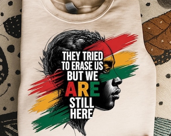 May include: Beige sweatshirt featuring a black and white portrait with red, yellow, and green brushstrokes. The text reads "THEY TRIED TO ERASE US BUT WE ARE STILL HERE."