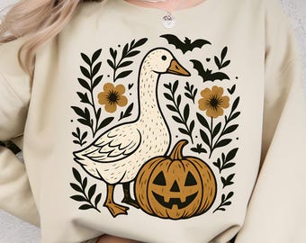 May include: Cream-colored sweatshirt featuring a cartoon goose, a jack-o'-lantern, and floral and bat graphics. The goose is white with a yellow beak and feet. The pumpkin is orange with a carved face. Black and brown accents.