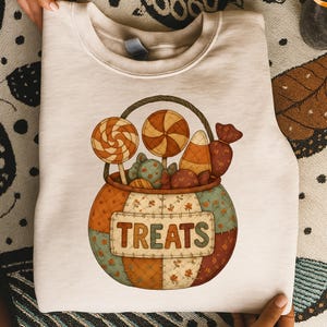 May include: A cream-colored sweatshirt featuring a colorful illustration of a basket filled with candies and lollipops. The basket has a handle and the word "TREATS" is displayed on the front. The design has a patchwork style.