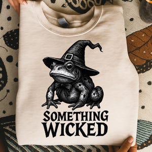 May include: A beige sweatshirt with a black and white illustration of a frog in a witch hat and bow tie. The words "SOMETHING WICKED" are printed below. The sweatshirt is made of a soft material, perfect for autumn.