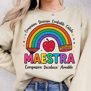 Puede incluir: Sudadera beige con un dise&ntilde;o de arco&iacute;ris colorido, una manzana roja con purpurina y la palabra "MAESTRA" en letras grandes y multicolores. El dise&ntilde;o incluye estrellas y palabras en espa&ntilde;ol.