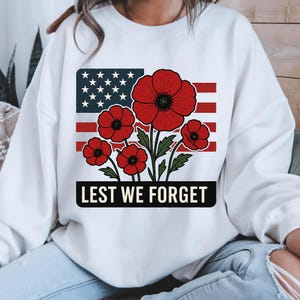 May include: White sweatshirt featuring a graphic of the American flag with red poppies and the text "Lest We Forget" in a black rectangle. The design includes red, white, blue, and green colors.