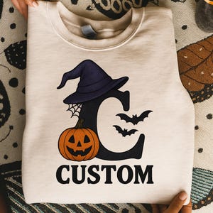 Puede incluir: Sudadera color crema con un diseño de Halloween. El diseño incluye la letra negra "C" con un sombrero de bruja, telaraña, calabaza y murciélagos. La palabra "CUSTOM" está impresa en negro debajo.