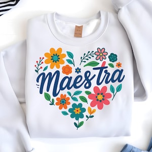 Puede incluir: Sudadera blanca con un dise&ntilde;o floral en forma de coraz&oacute;n. El coraz&oacute;n est&aacute; lleno de flores coloridas y hojas verdes, con la palabra "Maestra" en azul. Las flores son en tonos naranja, rosa y turquesa.