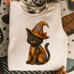 May include: A cream-colored sweatshirt featuring a stitched black cat wearing a patchwork witch hat with star accents and a bow. The cat has button eyes and a patchwork body in shades of orange, brown, and green. The design is centered on the front of the sweatshirt.