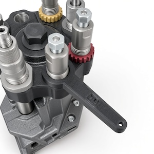 May include: A black 3D-printed tool with a handle and a circular opening, positioned around a silver metal component on a reloading press. The tool features a small embossed logo. The press includes multiple silver metal dies, a red adjustment ring, and a gold ring.