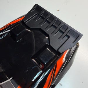 May include: A black and orange remote control car body shell featuring a matte black rear spoiler with a ribbed design. The glossy black surface is accented with bright orange graphic stripes and moulded geometric shapes across the top section.