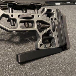 May include: A gray metal rifle stock assembly featuring an adjustable cheek riser, a rear butt pad, and a lower monopod attachment. The skeletonized frame includes multiple adjustment knobs, threaded screws, and black hardware components.