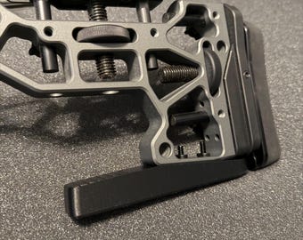 May include: A gray metal rifle stock assembly featuring an adjustable cheek riser, a rear butt pad, and a lower monopod attachment. The skeletonized frame includes multiple adjustment knobs, threaded screws, and black hardware components.