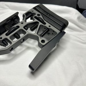 Op de afbeelding: Een zwart en grijs metalen geweerkolven met een zwarte rubberen achterplaat. De kolf heeft een skeletvormig ontwerp en een hendel aan de zijkant.