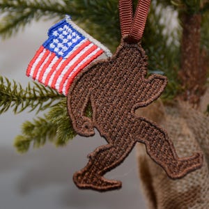 May include: Brown felt ornament of a Bigfoot holding an American flag. The ornament is hanging on a Christmas tree.