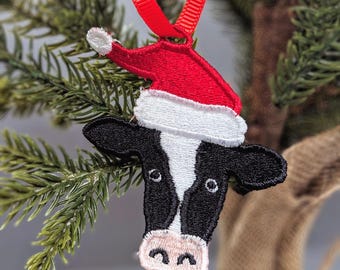May include: A black and white cow ornament wearing a red and white Santa hat. The ornament is hanging from a red ribbon on a Christmas tree.