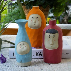 Può includere: Tre vasi in ceramica colorati con fantasiosi disegni di volti. I vasi sono nelle tonalità del blu, dell'arancione e del rosso. I volti hanno uno stile minimalista con tratti semplici. I vasi sono esposti su una superficie bianca con vegetazione sullo sfondo.