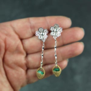 May include: A pair of silver-toned drop earrings featuring a five-petaled flower stud with intricate filigree detailing. A thin, wavy silver-toned metal wire connects the flower to a small, tan-colored woven basket charm containing a green element.