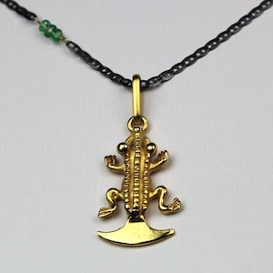 May include: A gold-toned lizard pendant suspended from a dark, thin chain. The lizard features a textured back and a wide, flat tail. A small cluster of three tiny green beads is threaded onto the dark chain to the left of the pendant bail.