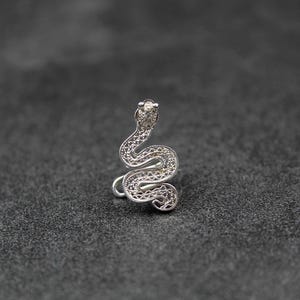 Puede incluir: Un pendiente tipo ear cuff de color plateado con forma de serpiente enroscada. El cuerpo de la serpiente presenta un intrincado patrón de filigrana calada con una superficie texturizada. La cabeza está ligeramente elevada y tiene una piedra clara.