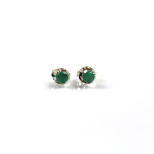May include: A pair of stud earrings featuring round, faceted green gemstones set in a polished silver-toned metal prong setting. The stones are a deep, vibrant green color and are secured by four small metal prongs on each earring base.