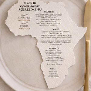 Op de afbeelding: Een crèmekleurige menukaart in de vorm van het continent Afrika, met zwarte tekst getiteld Black in Government Soirée Menu, met een lijst van voorgerechten, hoofdgerechten, desserts en bijgerechten, geplaatst op een rond keramisch bord met een goudkleurig mes.